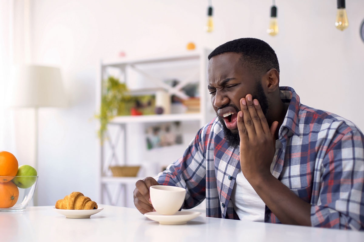 Man grimacing in sudden tooth pain while drinking morning coffee at home, holding jaw in discomfort