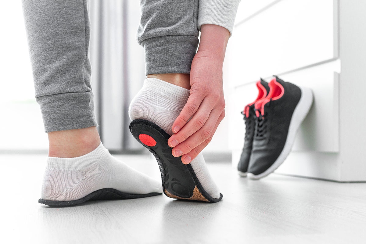 Orthotic insole examined from below, showing cork midsole and red heel pressure point, with running shoes in background