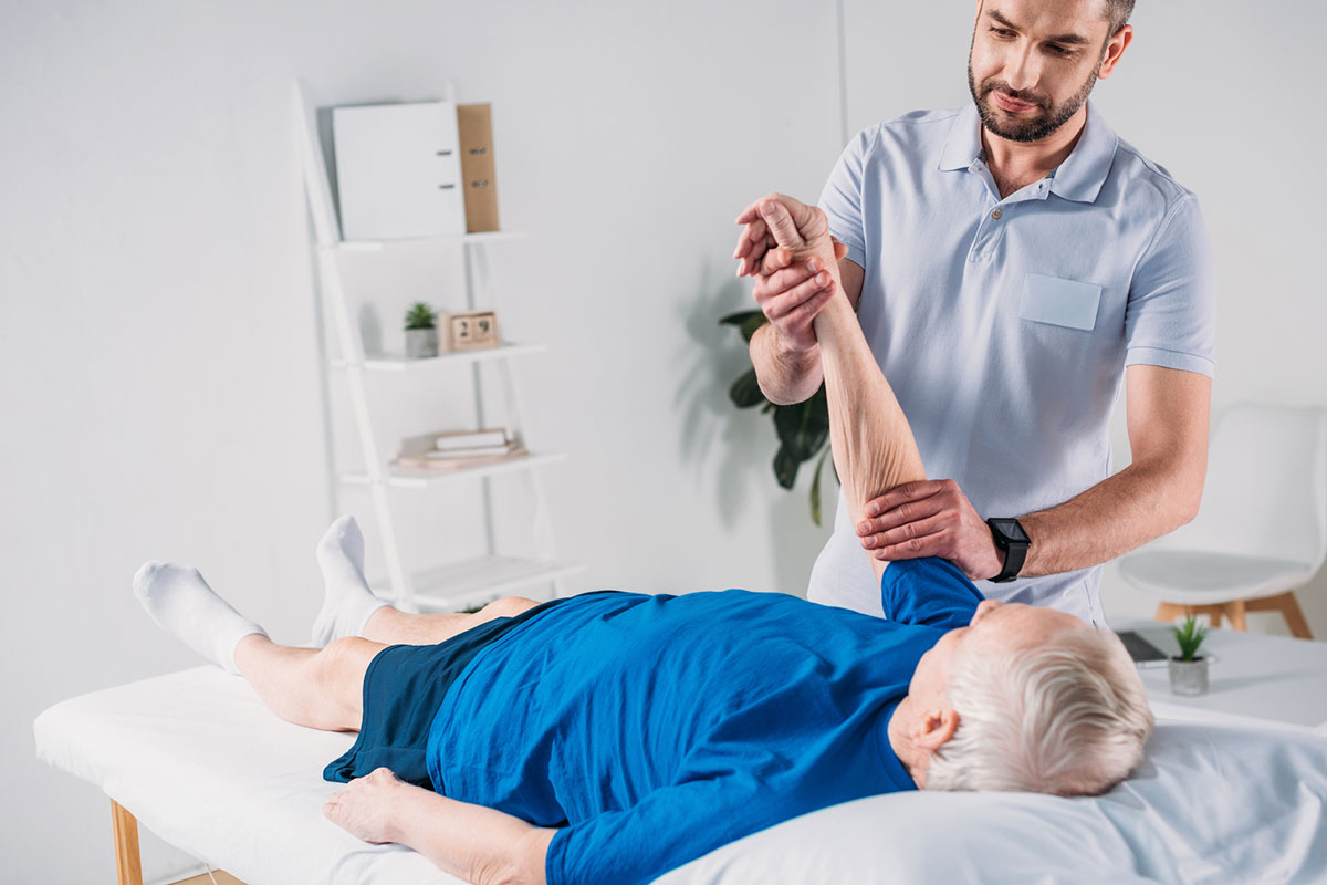Physiotherapist performing passive arm stretch on elderly patient lying on treatment table in modern clinic