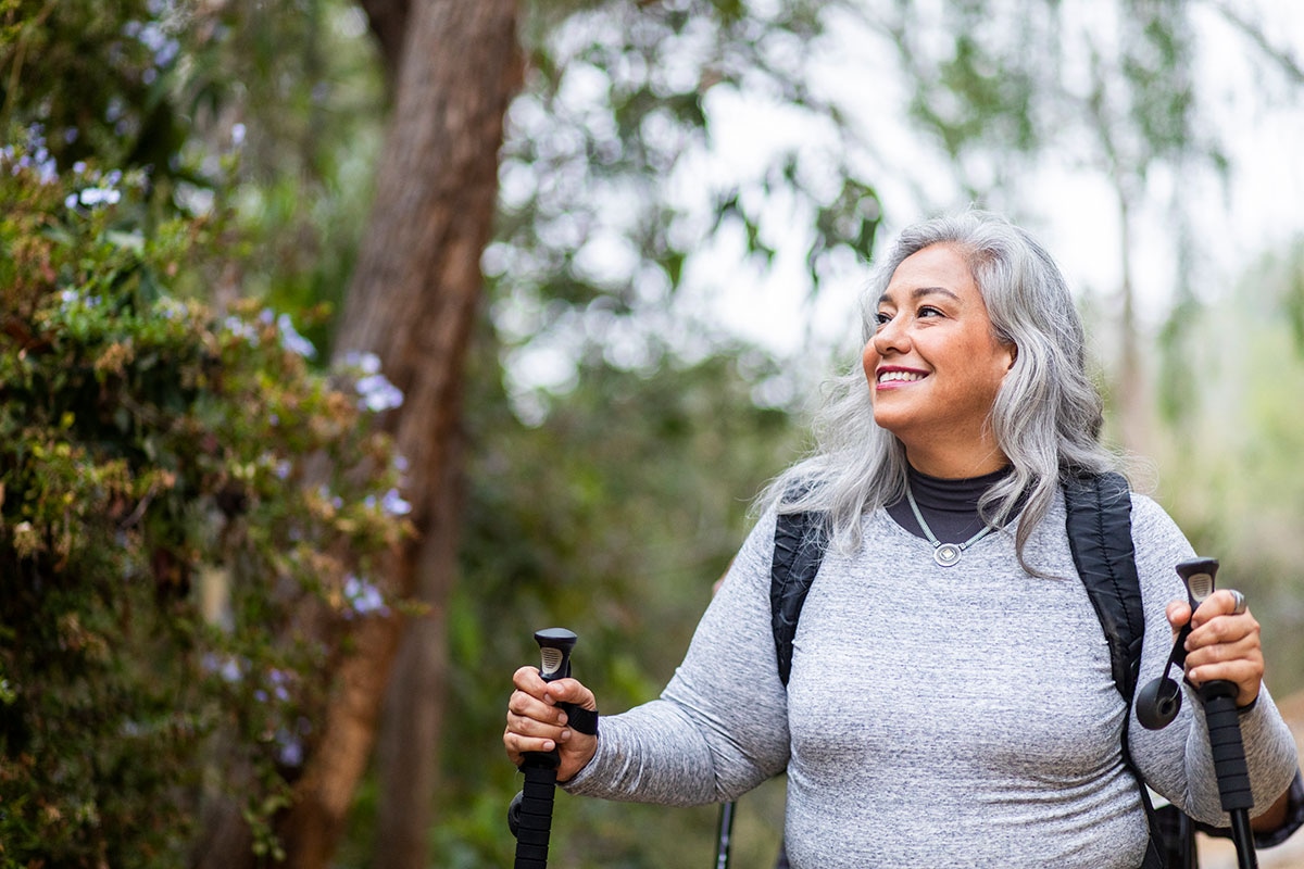Senior Hispanic woman hiking in a green forest with trekking poles and backpack, smiling confidently outdoors