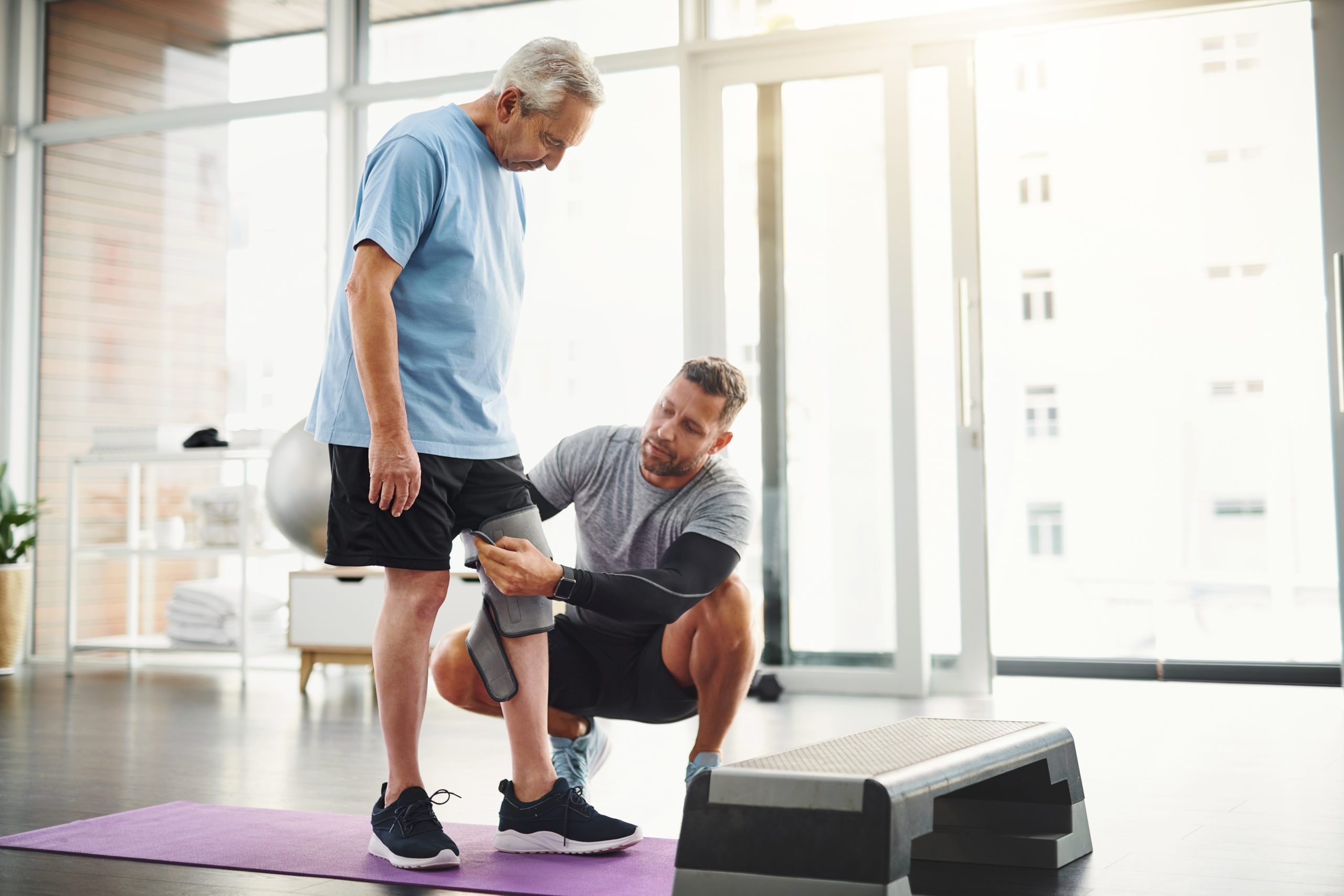 Senior patient working with physiotherapist on knee rehabilitation using step platform and yoga mat