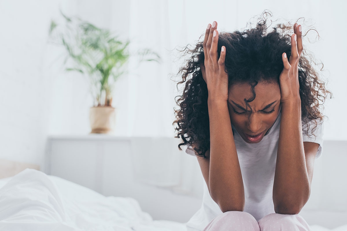 Woman sitting on bed pressing hands to head in pain from a severe migraine or headache